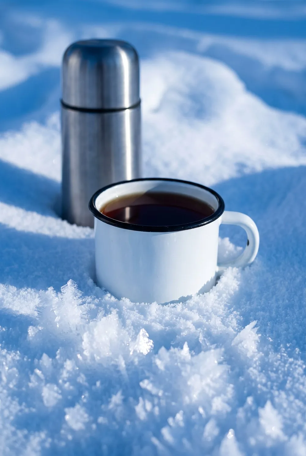 A white enamel mug filled to the rim with dark tea, set down into deep fresh snow with a steel vacuum flask behind it, huge sharp frost crystals in the foreground, the snow deep blue in shadow and brilliant white where the light catches it.