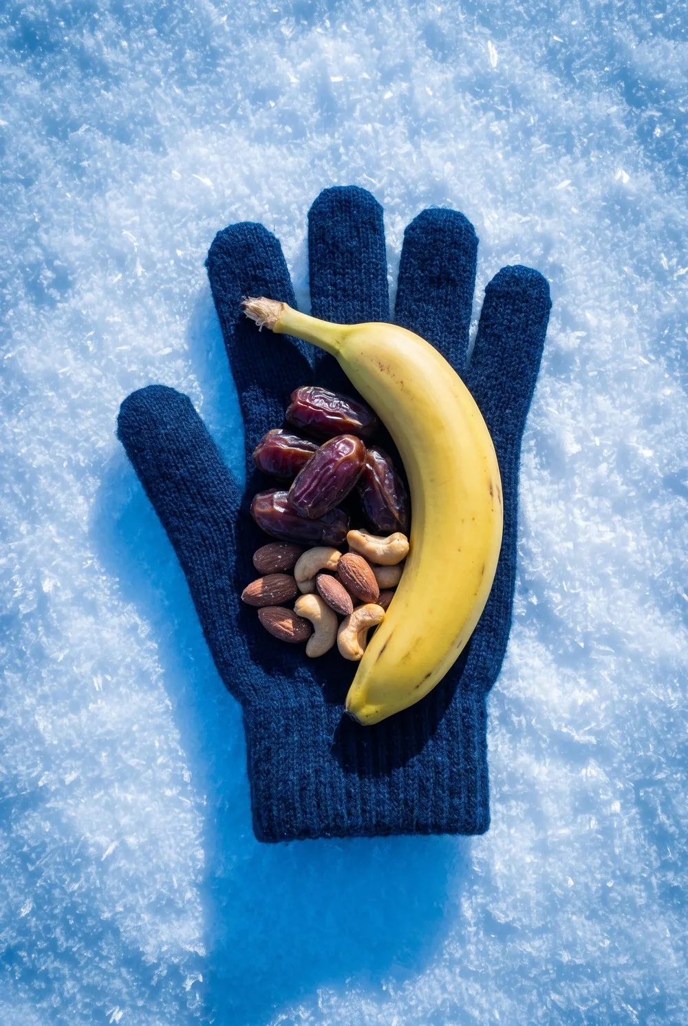 An overhead view of a plain knitted glove lying open on fresh snow with a whole banana, a pile of soft dates and a handful of salted nuts resting on it, sharp frost crystals scattered across the snow around it.
