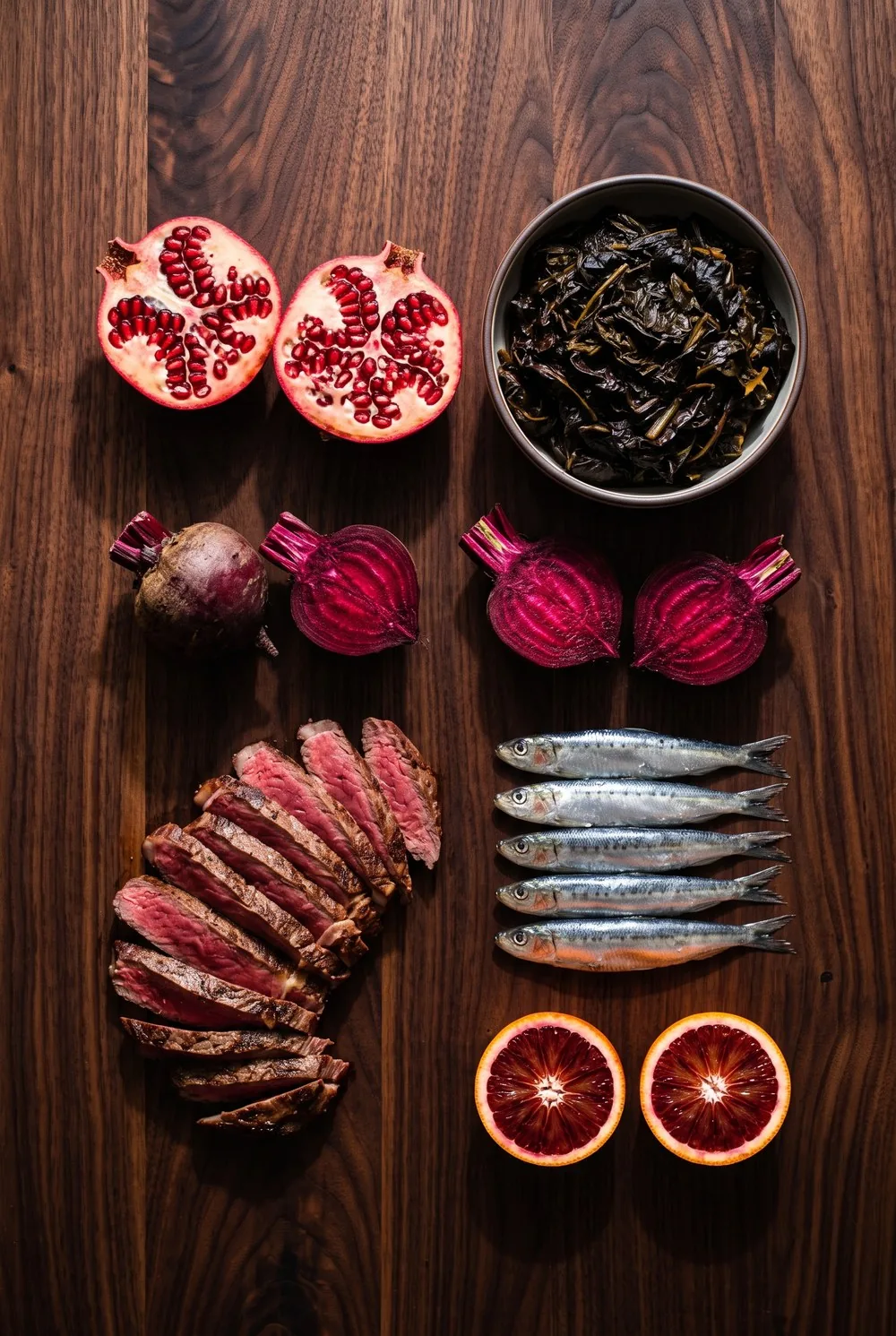 An overhead view of iron-rich foods arranged with clear space between them on dark walnut: two halved pomegranates seed side up, a bowl of dark cooked greens, roasted beets cut to show deep magenta rings, a fan of sliced rare steak, a row of silver sardines and two halved blood oranges, all deeply saturated against the warm dark wood.