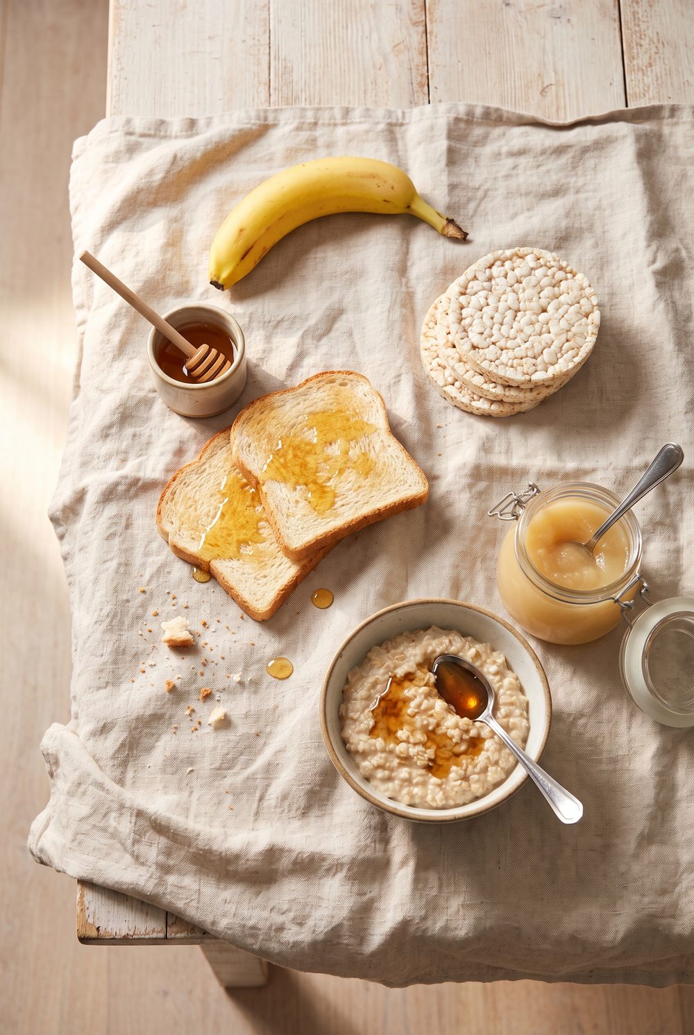 A pale wooden breakfast table in soft morning light with a banana, white toast with honey, plain rice cakes, a small bowl of oats with maple syrup and an open jar of applesauce arranged loosely on rumpled linen.