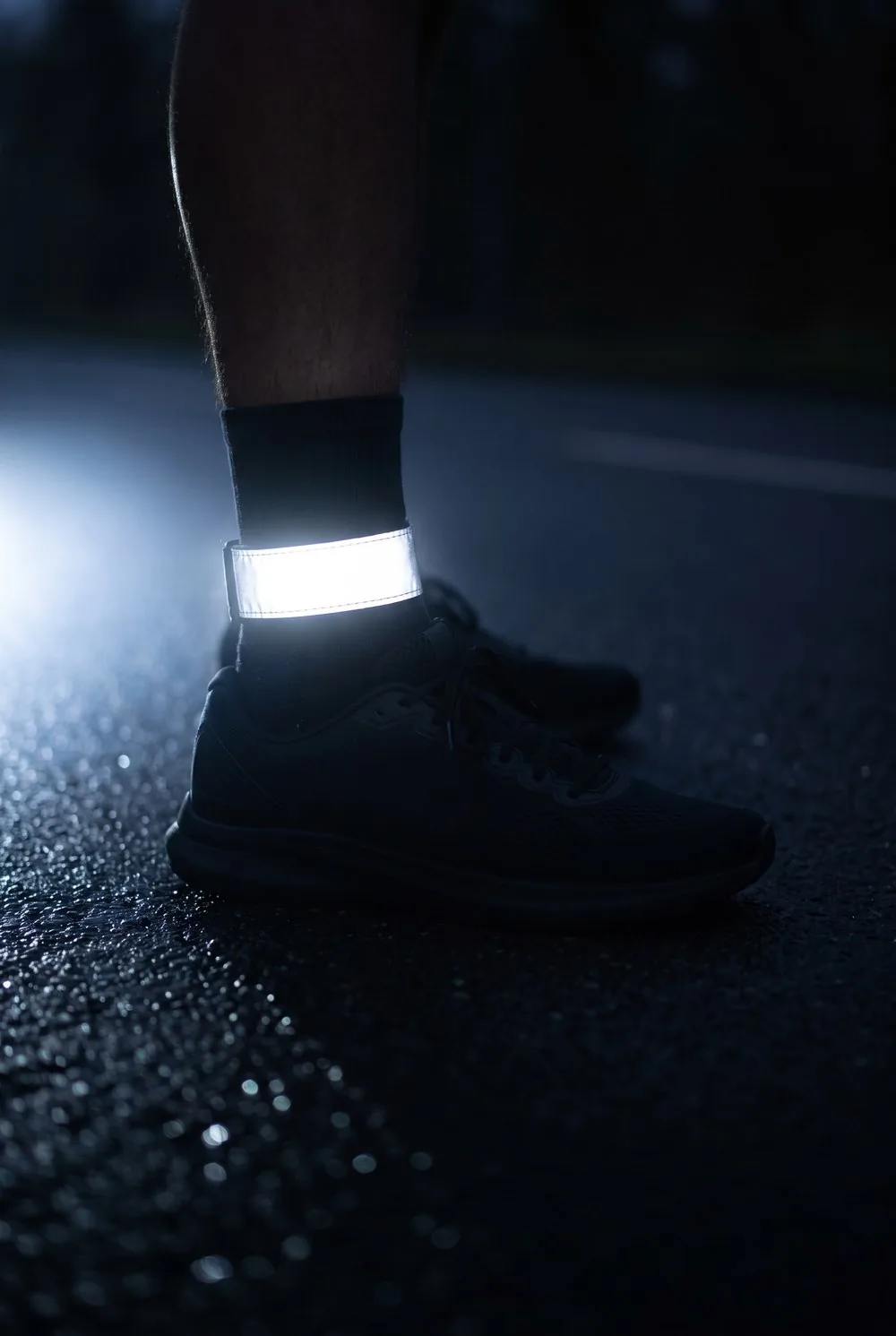 A tight close view of a reflective band fastened around a runner's ankle above a running shoe on a dark wet road before dawn, the reflective material blazing silver-white where a headlight catches it, everything around it falling to near black.