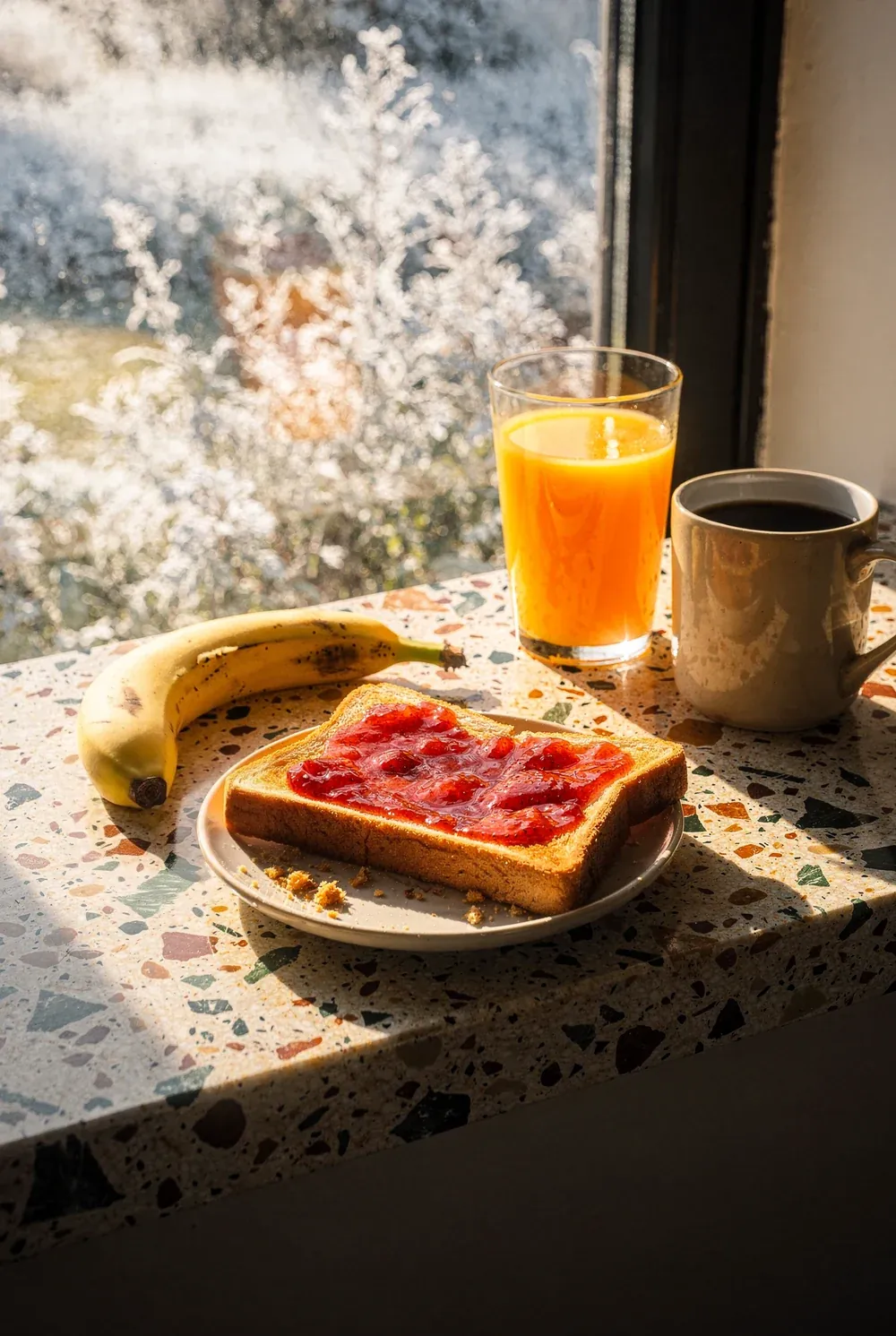 A thick slice of golden toast spread with strawberry jam on a cream plate with a few crumbs beside it, a whole banana, a full glass of orange juice glowing in the backlight and a mug of black coffee, set on a cream terrazzo ledge flecked with rust and green, low sun pouring through a frosted window behind them.
