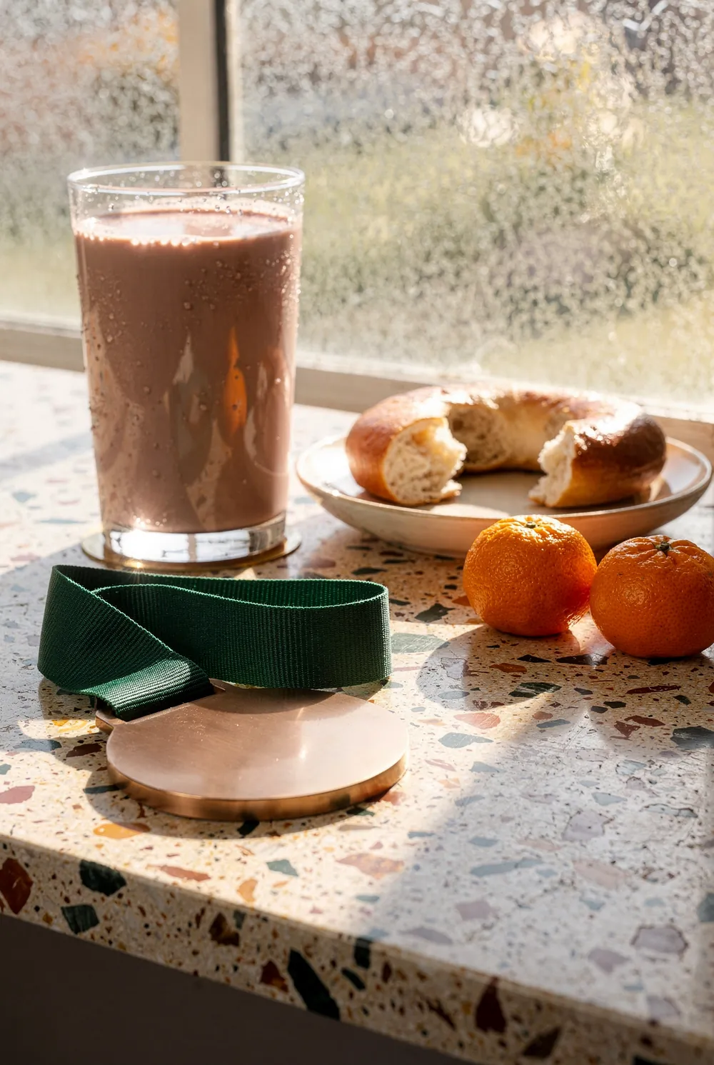 A plain bronze medal on a forest green ribbon lying on cream terrazzo in hard backlight, behind it a tall glass of chocolate milk beaded with condensation, a bagel torn in half on a cream plate and two clementines, a frosted window glowing behind them.