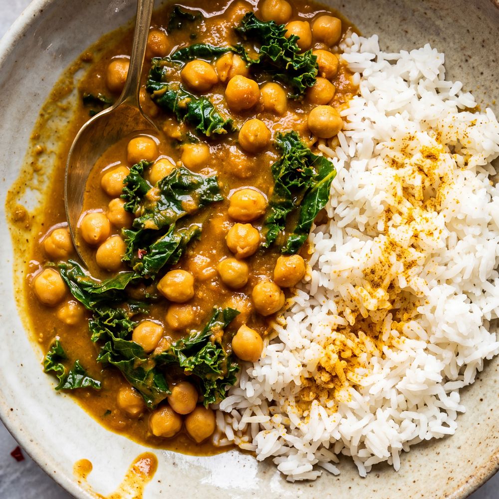 Overhead view of a golden coconut chickpea curry in a wide oat-colored stoneware bowl, whole chickpeas piled thick in the sauce with dark torn kale stirred through, a mound of steamed white rice beside it, gold spoon on a white marble surface.