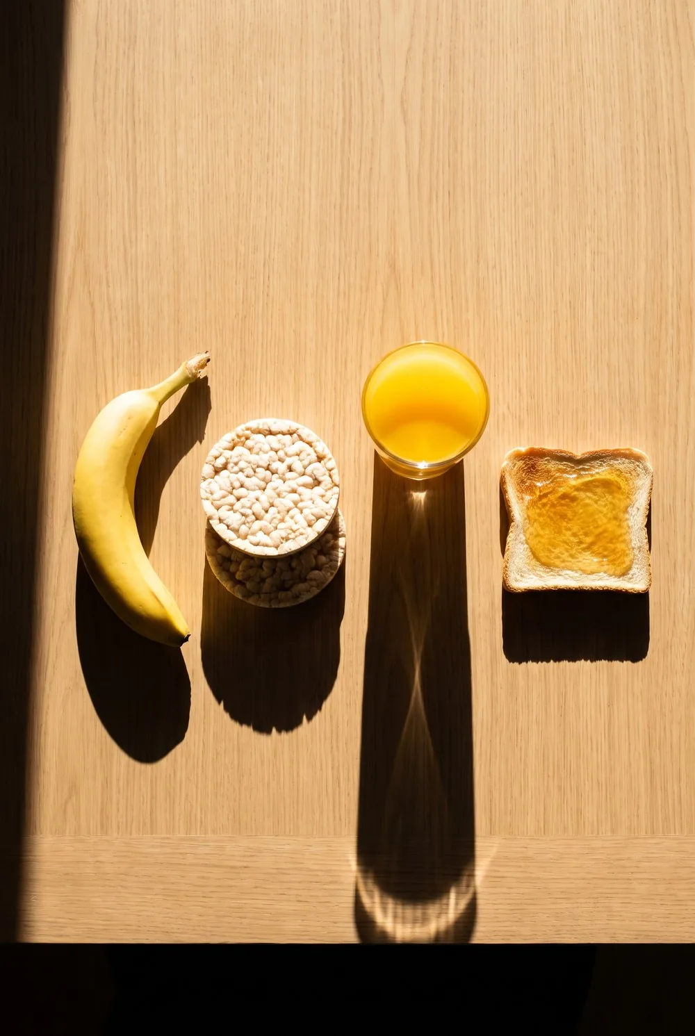 An overhead view of a pale oak kitchen counter in hard low morning sun, with a banana, two plain rice cakes, a small glass of orange juice and a slice of white toast with honey set well apart on the bare wood, each throwing a long crisp shadow across the grain.