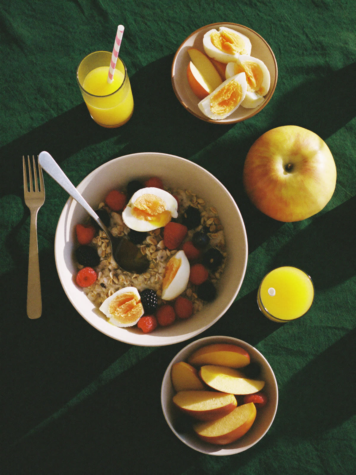 A generous breakfast on a deep green cloth: oats with berries and soft eggs, apples, and orange juice in morning sun