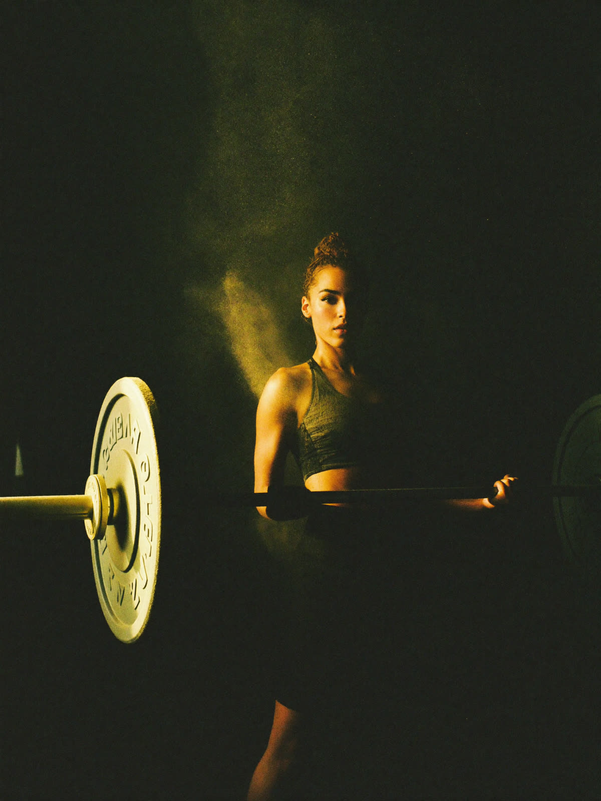 A female athlete holding a loaded barbell in a dark gym, lit by a single warm beam of light